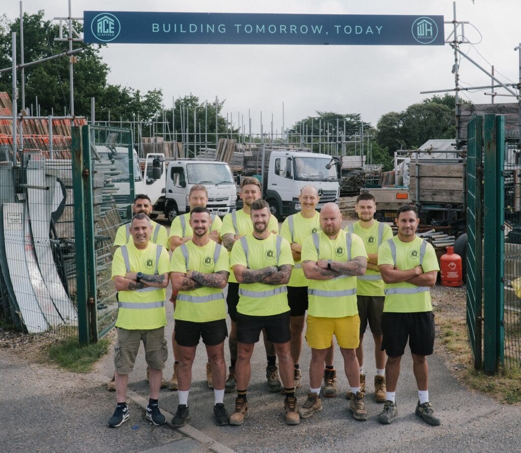 A team of ten scaffolders wearing bright yellow ACE Scaffold high-visibility shirts posing together at a scaffolding yard gate, under a "Building Tomorrow, Today" sign, with work trucks parked behind them.