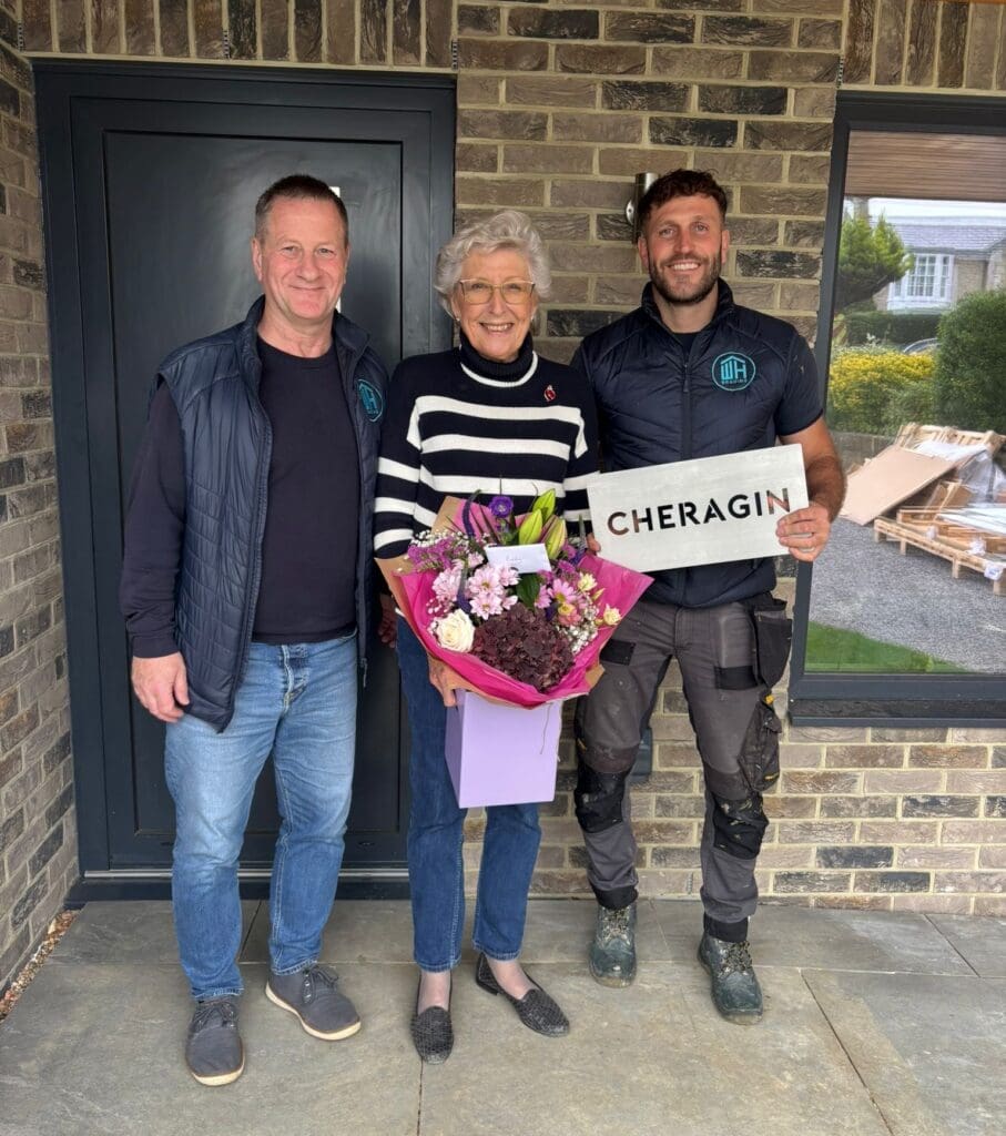 Three people smiling outside a modern front door. The person in the center holds a large bouquet of flowers, and the person on the right, wearing a "WH BRADING" work vest, holds a metal "CHERAGIN" house sign.