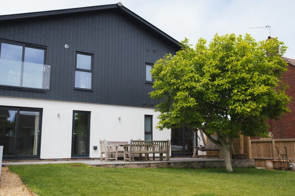 Modern home rear exterior featuring a stark contrast of dark grey vertical siding above white render, with large glass doors opening onto a patio with a wooden dining set next to a leafy tree.