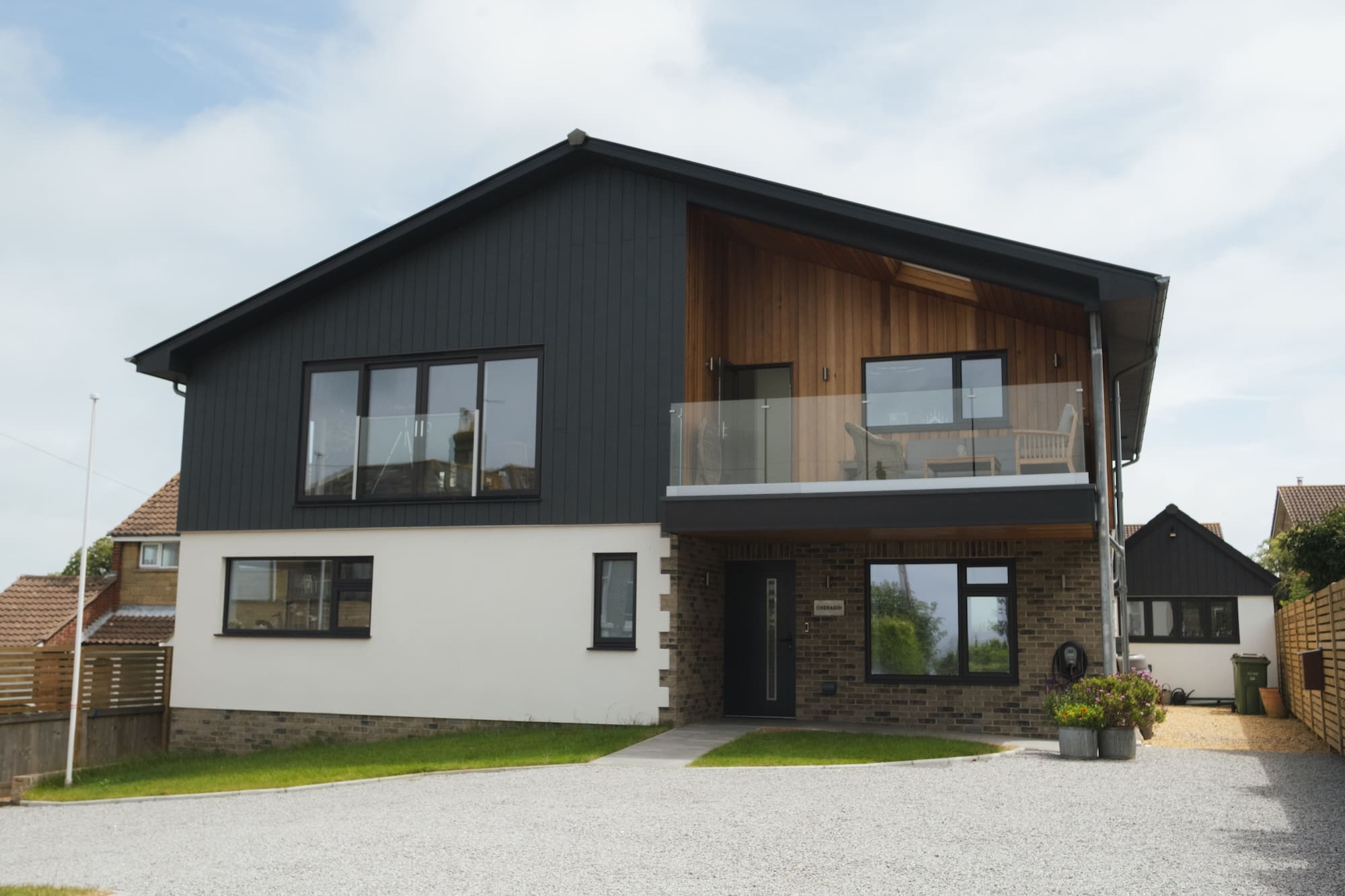 Wide front exterior view of a modern detached house with dark grey cladding, a contrasting natural wood balcony area, large contemporary windows, and a gravel driveway.