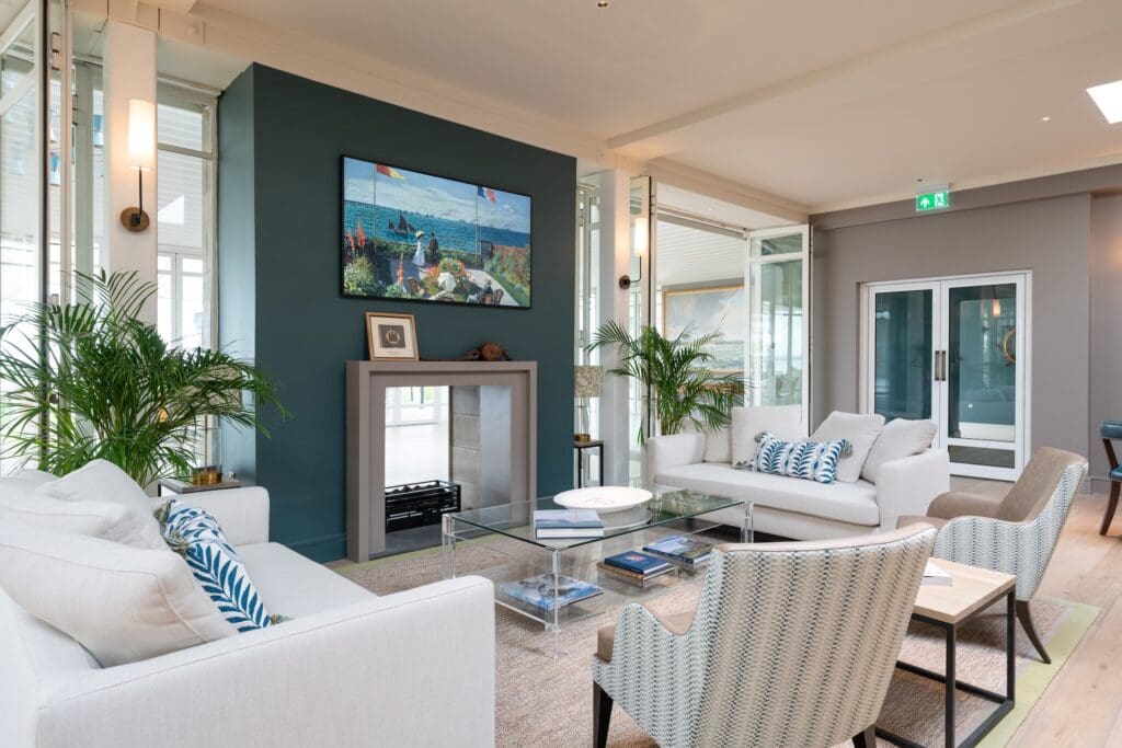 Modern living room with two white sofas and patterned chairs around a glass coffee table. A framed seascape painting hangs above a sleek fireplace.