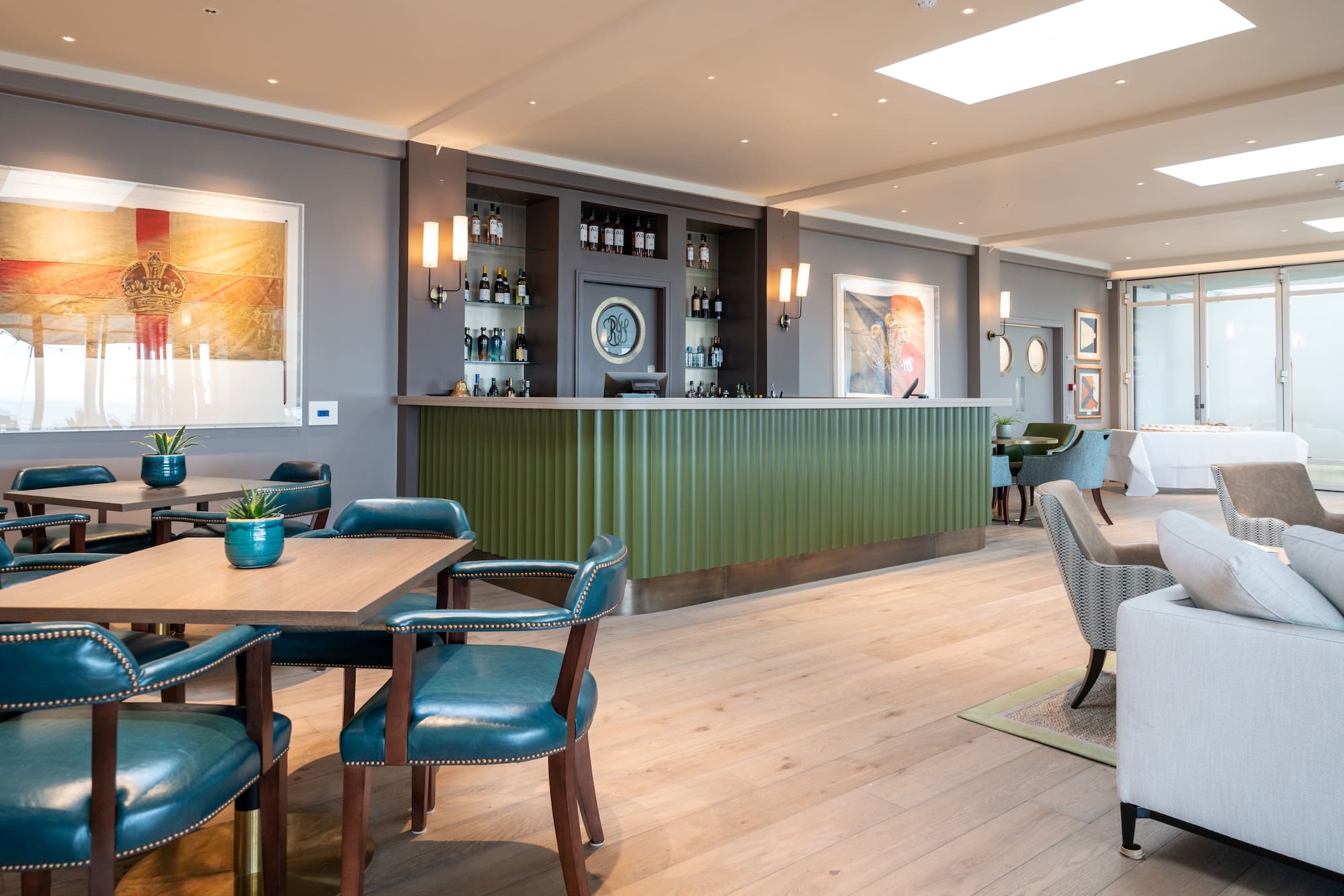 Wide view of a bright bar and lounge area showing blue leather seating, a green bar counter, and a large framed vintage flag artwork on the wall.